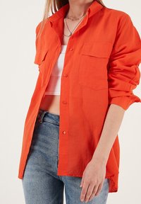 Femme portant une chemise orange vif à boutons sur un débardeur blanc court et un jean bleu, avec des colliers superposés et des manches retroussées.