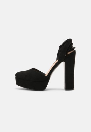 Escarpin plateforme en suède noir avec un bout arrondi, un dos ouvert et un détail de volants floraux au niveau du talon. Talon bloc transparent.