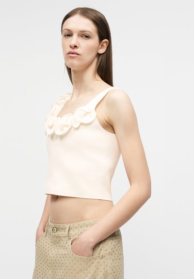 Γυναίκα με ίσια καστανά μαλλιά που φοράει ροζ παστέλ κοντό τοπ χωρίς μανίκια με υφασμάτινες λουλούδι λεπτομέρειες και μπεζ παντελόνι με τρουκς, έχοντας τα χέρια στις τσέπες.