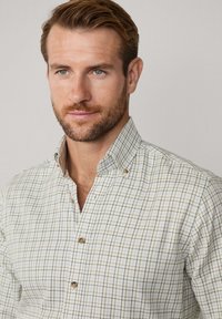 Man met lichtbruin haar en een baard, gekleed in een beige geruit overhemd, kijkt iets naar rechts tegen een neutrale achtergrond.