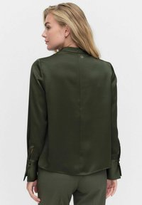 Tummanvihreä satiinipaita, jossa on korkea kaulus, pitkät hihat napitetuilla hihoilla ja avaus yksityiskohtana selässä. Sujuva tekstuuri.