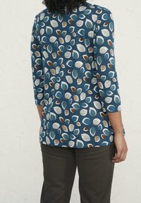 Top a maniche lunghe blu con un motivo di foglie multicolore; realizzato in tessuto morbido con una vestibilità ampia. Abbinato a pantaloni marroni scuri.