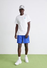 Vit bomullströja med turkos Nike-logga, blå shorts, vita sneakers och en grå keps med logga, stående på en grön yta.