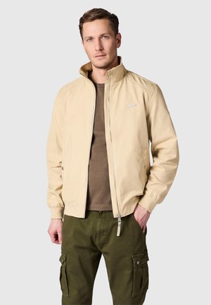 Homme portant une veste beige zippée sur une chemise marron et un pantalon cargo vert olive, debout avec une main dans la poche sur fond uni.