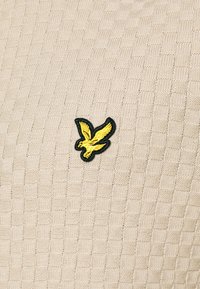 Gelbes besticktes Vogel-Logo auf beigem strukturiertem Strickstoff mit quadratischem Muster.