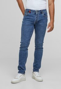 Kék farmernadrág slim fit szabással, öt zsebes dizájnnal, piros díszítéssel a derékszíjon, és enyhe kopással. Fehér sportcipővel viselve.