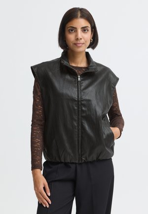 Femme aux cheveux courts et foncés porte une veste en cuir noire sans manches sur un haut en dentelle marron et un pantalon noir, debout devant un fond uni.