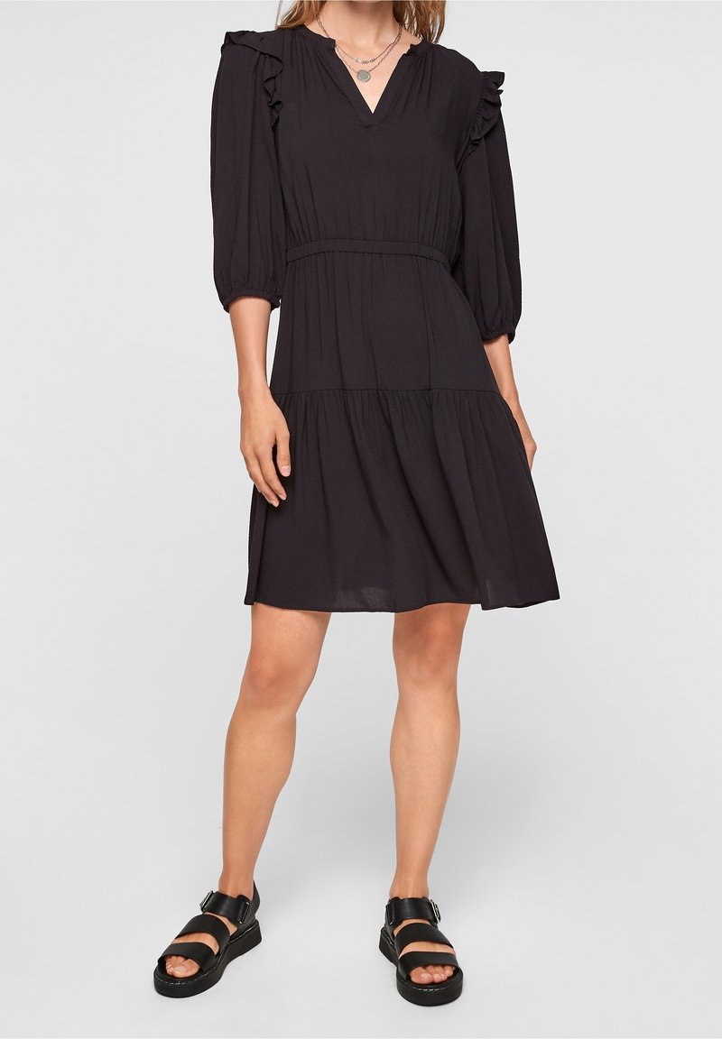 Femme portant une robe noire longueur genou avec des épaules volantées et des manches trois quarts, associée à des sandales noires à semelles épaisses avec des boucles.