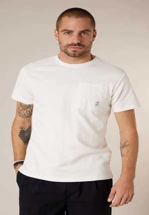 Homme aux cheveux courts et à la barbe portant un t-shirt blanc avec une poche et un pantalon noir, montrant des tatouages sur les deux avant-bras et des bracelets au poignet gauche.