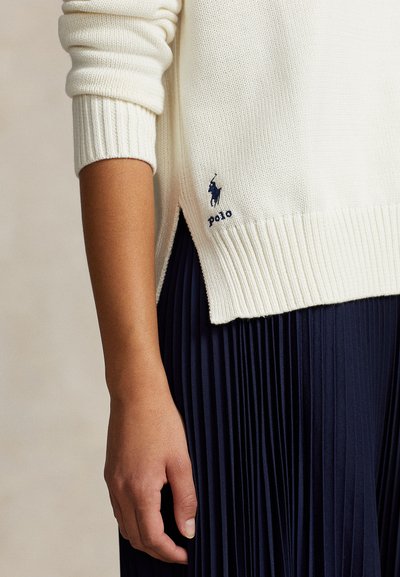 Kremiškai baltas megztinis su ribinėmis rankogaliais ir apačia, šone su tamsiai mėlynu logotipu, derinamas su tamsiai mėlyna klostuota sijonu.