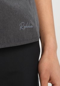 Gros plan sur la main d'une personne à côté d'un tissu gris avec le logo "Rukka" au-dessus d'un vêtement noir.