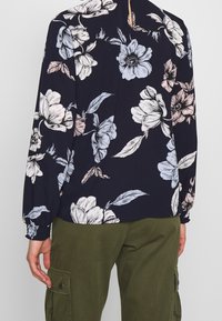 Blumenbluse in dunklem Marineblau mit hellroten und blauen Blumen. Lange Ärmel mit elastischen Bündchen; Schlüsselband-Detail im Rücken. Kombiniert mit olivgrünen Hosen.