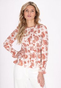 Blusa floreale a maniche lunghe in un tessuto morbido, con una parte superiore arricciata e polsini a balze, su uno sfondo crema chiaro con motivi color ruggine.