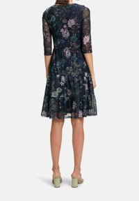 Robe à motif floral avec un fond bleu marine, manches en dentelle trois-quarts, jupe à volants et texture transparente. Associée à des mules à talons carrés verts.