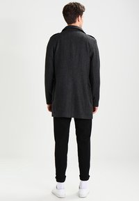 Abrigo de lana gris oscuro con un corte largo, cuello de muesca, bolsillos laterales y hombreras, combinado con pantalones negros y zapatos blancos.