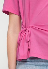 Rosa Kurzarm-Top mit seitlicher Knoten-Detail. Das aus weichem Stoff gefertigte Top zeichnet sich durch eine lockere Passform und einen mehrlagigen Saum mit glatter Textur aus.