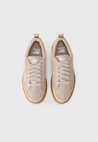 Et par beige semskede sneakers med tykke brune såler og hvite snørebånd, sett ovenfra på en hvit bakgrunn.