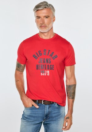 Mężczyzna z siwymi włosami i brodą, ubrany w czerwoną koszulkę z napisem "Big Star Jeans Heritage 1973" oraz niebieskie dżinsy, stoi z jedną ręką w kieszeni.