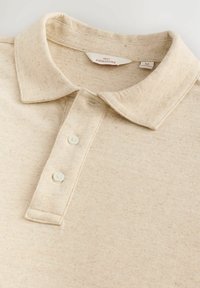 Beige polo skjorte med et tekstureret stof, der har en klassisk krave, tre knapper og subtile prikker i materialet.