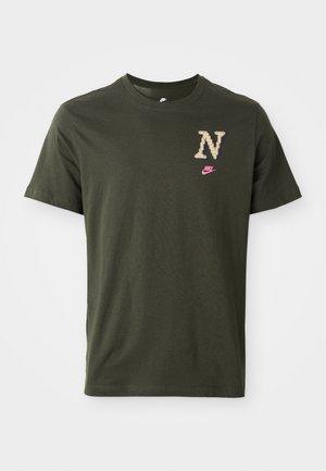 Olivnozeleni bombažni majica s kratkimi rokavi, ki ima velik vzorčen logotip "N" v bež barvi in majhen roza Nike znak na prsih.