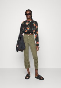 Top preto de mangas longas com padrão floral, calças cropped de cintura alta em verde-azeitona com bordados de flores, e sandálias pretas tipo slide. Casaco pousado sobre o braço.