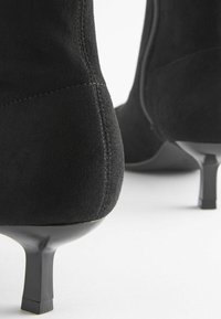 Czarne zamszowe botki na kostkę z zwężającym się obcasem, charakteryzujące się gładką teksturą i minimalnymi detalami przeszyć, skupione na smukłej sylwetce.