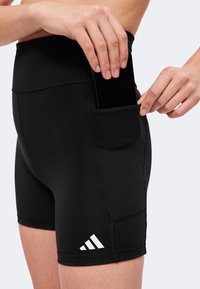 Hände legen ein schwarzes Smartphone in die Seitentasche einer schwarzen Sportshorts mit einem weißen Logo nahe dem Saum.