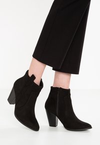 Botines de ante negro, con cremallera lateral y tacones cuadrados, llevados con pantalones negros recortados sobre un fondo claro.