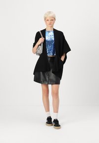 Svart stickad cardigan med öppen front över en blå paljett crop-top, matchad med en svart läder mini kjol med framtida knappar, och svarta sneakers.