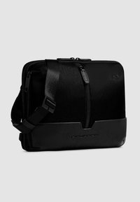 Schwarze Umhängetasche aus Textil und Leder, mit Reißverschluss, verstellbarem Trageriemen und geprägt Logo auf der Vorderseite. Kompakte rechteckige Form.