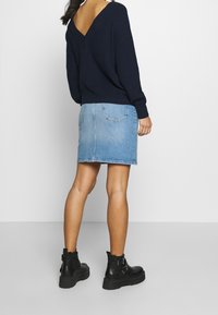 Marineblauer Strickpullover mit V-Ausschnitt, kombiniert mit einem hellblauen Jeanrock. Schwarze Stiefeletten mit Schnallenakzenten und strukturierten Sohlen.