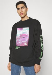 Čierne tričko s dlhým rukávom so živým grafickým potlačou v ružových a zelených farbách; text "Calvin Klein Jeans" v neónovo zelenej na rukáve.
