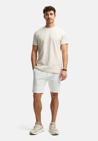 Beige short-sleeve t-shirt paired with white shorts and beige athletic shoes. The model stands with hands in pockets, neutral background.