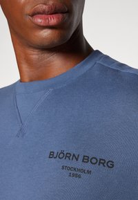 Sweatshirt azul com decote redondo, texto preto icónico "BJÖRN BORG STOCKHOLM 1956" e detalhes de costura visíveis.