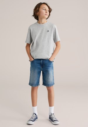 Szürke rövid ujjú póló kis fekete logóval, világoskék farmer rövidnadrággal és fehér bokazoknival, tengerészkék edzőcipővel viselve.