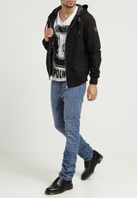 Czarna bluza z kapturem na zamek z troczkami, biały t-shirt z grafiką, niebieskie dopasowane jeansy z przetarciami oraz czarne buty.