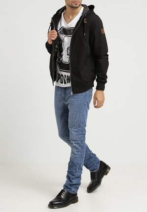 Czarna bluza z kapturem na zamek z troczkami, biały t-shirt z grafiką, niebieskie dopasowane jeansy z przetarciami oraz czarne buty.