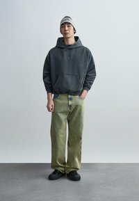 Σκούρα γκρι φούτερ με κουκούλα και τσέπη μπροστά, παντελόνι φαρδύ με κάθετες ανοιχτό πράσινες ρίγες, και μαύρα αθλητικά παπούτσια. Σκουφί με λεπτομέρεια λογότυπου.