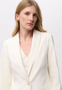 Krémový blazer s ostrým klopem, zapínání na jeden knoflík a přiléhavý střih. Nosený přes světle béžový top s výstřihem do V, vyrobený z hladké látky s jemným leskem.