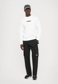 Felpa bianca con un'etichetta nera, pantaloni cargo neri con tasche e scarpe da ginnastica nere. Il cappellino è grigio con una fascia a fantasia.