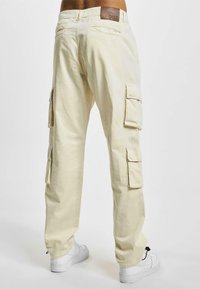 Pantalones cargo beige hechos de tela ligera, con múltiples bolsillos laterales y un parche de cuero en la cintura. Combinados con zapatillas blancas.