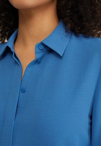 Camicia blu con colletto a polo, caratterizzata da due bottoni al colletto e da una texture di tessuto liscia e leggera.