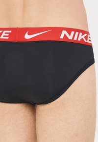 Black cotton briefs featuring a red elastic waistband with white "NIKE" logo and swoosh. Smooth texture, fitted design, and low-rise cut.