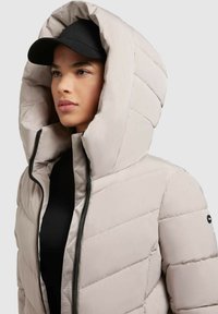 Lichtbeige pufferjack met een hoge, gewatteerde kraag, voorzien van een zwarte rits en een zijlogo patch. Gemaakt met een gequiltde textuur.