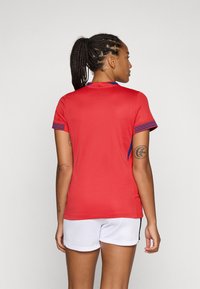 T-shirt de sport rouge à manches courtes avec des accents décoratifs bleus et à motifs, associé à un short blanc. La texture semble lisse.