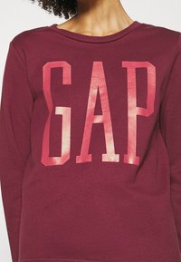 Sudadera de manga larga burdeos con un gran logo rosa "GAP" en el frente. Tejido suave y texturizado con cuello tipo crew.