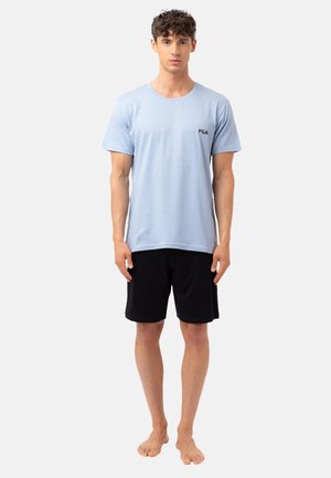 Jeune homme debout pieds nus, portant un t-shirt FILA bleu clair et un short noir, sur un fond blanc uni.