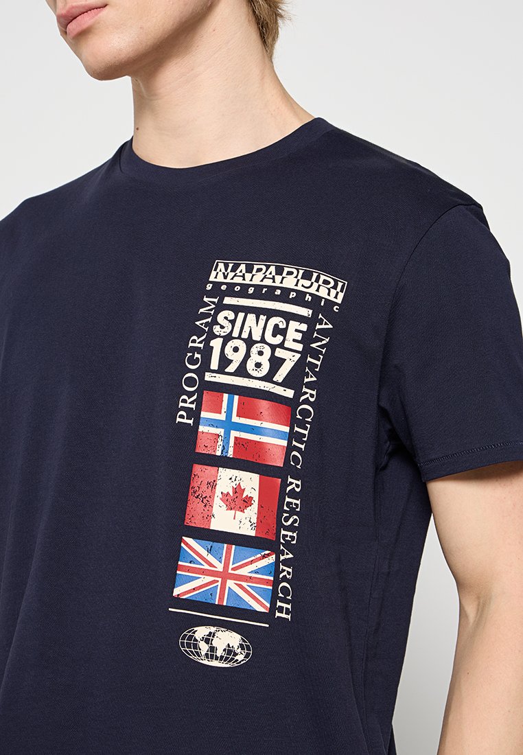 Mężczyzna w granatowej koszulce Napapijri z napisem "Od 1987 roku Program Badań Antarktycznych" oraz flagami Norwegii, Kanady i Wielkiej Brytanii.