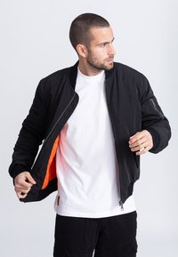 Zwarte bomberjack met ritssluiting, geribbelde manchetten en een oranje voering, gedragen over een wit T-shirt. Voorzien van een zijzak.