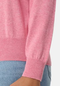 Růžový svetr s žebrovaným lemem a manžetami, vyrobený z měkké, texturované tkaniny. Detailní záběr ukazuje detail rukávu a hladký pletený vzor.